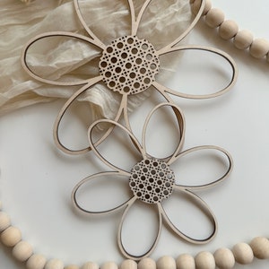 May include: Two wooden flower decorations with a natural wood finish. The flower petals are outlined in black, and the center features a woven cane design. The flowers are placed on a white surface with a beige fabric and wooden beads in the background.