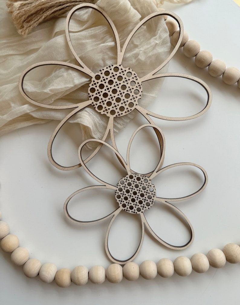 May include: Two wooden flower decorations with a natural wood finish. The flower petals are outlined in black, and the center features a woven cane design. The flowers are placed on a white surface with a beige fabric and wooden beads in the background.