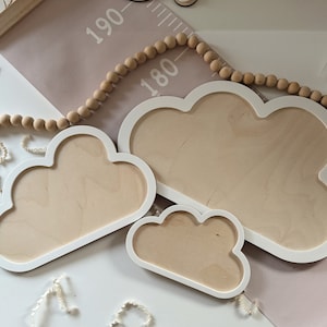 May include: Three cloud-shaped wooden frames with white borders and light wood interiors. A wooden bead garland and a pink and white height chart are in the background. The frames are different sizes.