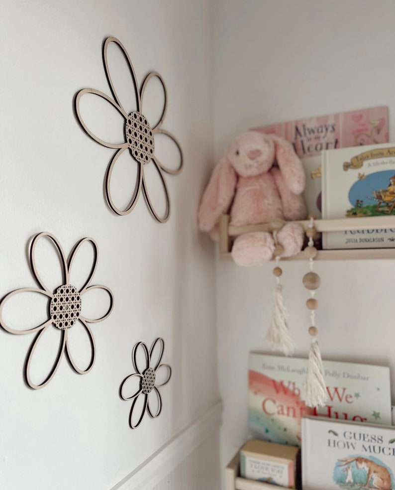 May include: Three wooden flower wall decorations with a natural wood finish and woven cane centers are mounted on a white wall. A pink stuffed bunny sits on a shelf with books. The flowers vary in size.