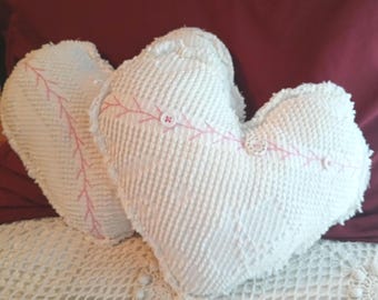 May include: Two heart-shaped pillows made of white textured fabric. Each pillow features a decorative pink stitched line running across the center, accented with small white buttons placed along the stitching to create a baseball-like design.