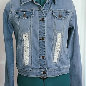 May include: A light blue denim jacket with white lace panels on the front. The jacket has a button closure and two front pockets.