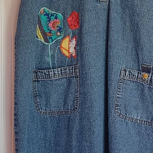 May include: A blue denim dress with a button-up collar and two patch pockets. The dress has a floral design on the front, made from colorful fabric scraps.