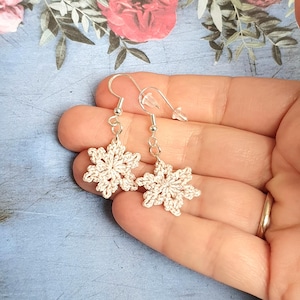 May include: A pair of snowflake earrings. The earrings are crafted from a light beige crocheted yarn and suspended from silver-tone hooks. The snowflake design is detailed, with multiple points radiating from the centre, creating a delicate appearance.