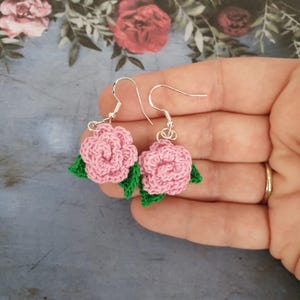 May include: A pair of handmade crochet earrings featuring pink rose-shaped designs with green leaves. The earrings have silver-coloured hooks. The background has a floral pattern.