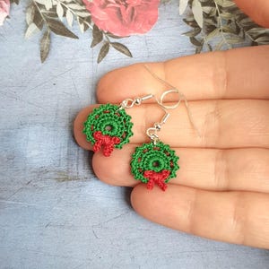 May include: Handmade crochet earrings. Each earring features a green wreath with a red bow. The earrings have silver-coloured hooks. These festive earrings are a charming accessory, ideal for the holiday season.
