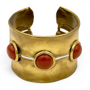 May include: A wide cuff bracelet made of brushed gold-tone metal featuring a split design. Three smooth, round, orange-red cabochon stones are set horizontally across the center of the band, with one in the middle and one on each side.