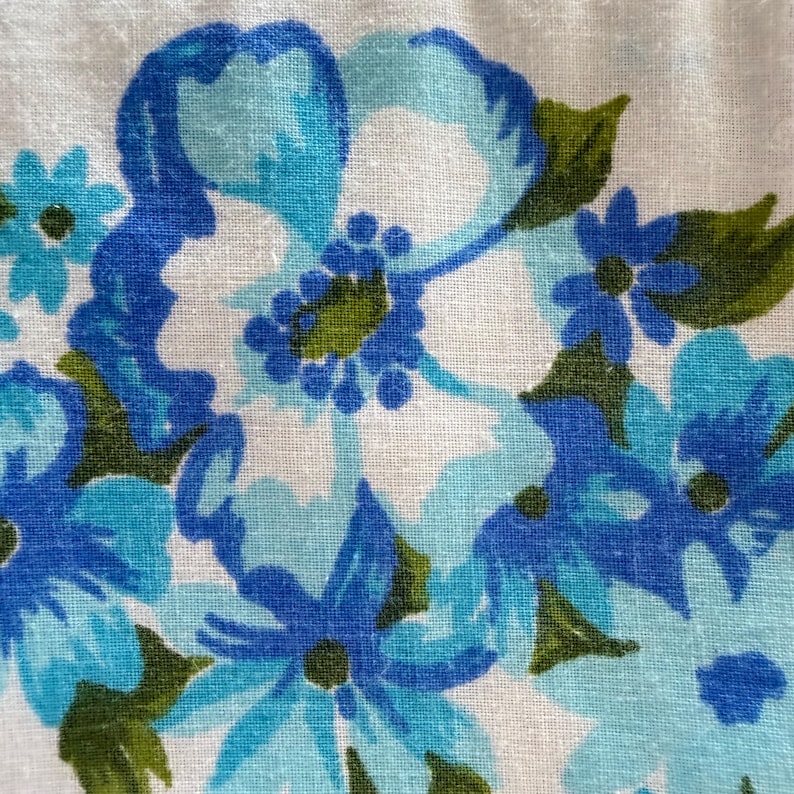 May include: Close-up of a fabric with a floral pattern. The design features blue and turquoise flowers with green leaves against a white background. The flowers have a vintage, hand-painted appearance, creating a soft, textured look.
