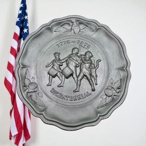 May include: A pewter-colored plate with a scalloped edge, featuring a raised relief design commemorating the Bicentennial. The design includes figures playing instruments and the text "1776-1976 Bicentennial". An American flag hangs in the background.