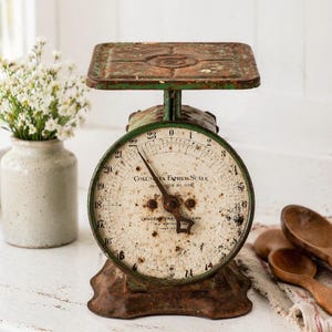 May include: An antique Columbia Family Scale with a distressed green and brown finish. The scale features a round face with a white background, black numerals, and a needle pointing to the weight in ounces. The scale has a rectangular top and a decorative base.