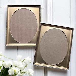 May include: Two gold-colored picture frames with oval openings. The frames have a metallic finish and a decorative border. The background is a white wooden surface. A bouquet of white flowers is in the lower left corner.
