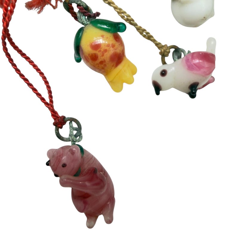 May include: A collection of glass charms hanging from colorful cords. The charms include a pink bear, a yellow and orange fruit-shaped piece with green leaves, and a white bird with pink accents, all attached by small metal loops to red and gold cords.