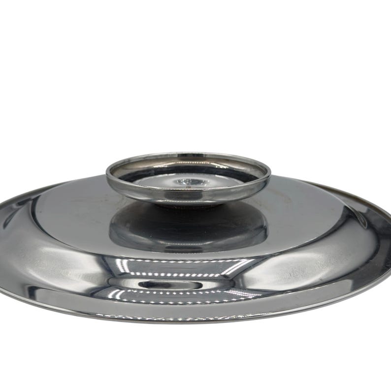 Op de afbeelding: Een cirkelvormig, gepolijst zilverkleurig metalen deksel met een glad, reflecterend oppervlak en een centrale, verhoogde ronde handgreepknop. Het deksel heeft een licht gewelfd profiel met een strakke, metalen afwerking die het licht weerkaatst.