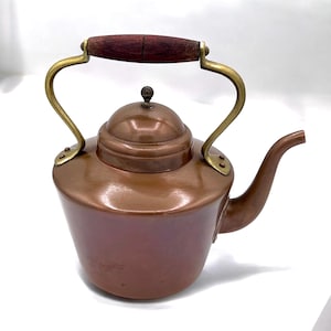 Puede incluir: Una tetera de cobre con un asa de madera y detalles de latón. La tetera tiene un cuerpo redondeado y un pico en el lateral.