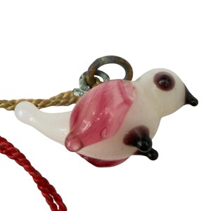May include: A glass bird pendant featuring a translucent white body with pink wing accents and dark purple details on the eyes and feet. The bird is attached to a weathered metal jump ring and is suspended by a red and gold braided cord.