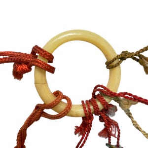 May include: A smooth, off-white circular ring made of a hard material, featuring a visible hairline crack. Multiple strands of red, orange, and brownish-green braided cord are knotted and wrapped securely around the circumference of the ring.