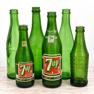 Puede incluir: Una colección de seis botellas de refresco vintage de vidrio verde. Dos botellas tienen etiquetas 7up rojas y blancas con el texto Fresh Up with 7up y You Like It It Likes You. Las otras cuatro son de vidrio verde, una con patrón de puntos.