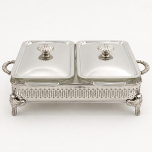 May include: A silver-plated double buffet server featuring two rectangular glass inserts with matching silver lids. The base has an intricate pierced lattice design, four ornate feet, and two side handles with a polished metallic finish.