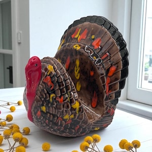 Puede incluir: Una figura de pavo de cerámica con cabeza y carúncula rojas, cuerpo marrón y abanico de plumas en tonos marrones, negros, amarillos y naranjas. El pavo es un objeto decorativo, probablemente para Acción de Gracias o decoración otoñal.