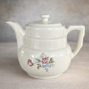 Puede incluir: Tetera de cerámica color crema con diseño floral. La tetera tiene un cuerpo redondeado, un pico curvo y un asa. El diseño floral incluye flores rosas, moradas y azules con hojas verdes. La tapa tiene un pequeño pomo.