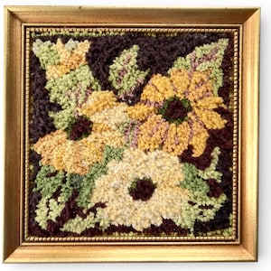 以下が含まれることがあります： 額装された正方形のフローラルテキスタイルアート作品。このアートワークは、濃い茶色の背景に、黄色、クリーム色、緑色の花のブーケを特徴としています。フレームは金色です。