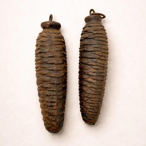 May include: Two antique, brown clock weights with a textured, cone-like shape. Each weight has a small loop at the top. The weights are made of a dark, possibly metal material, and show signs of age and wear.