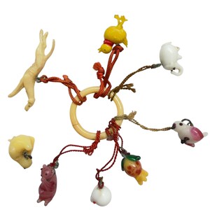 May include: A central cream-colored ring holds eight small charms attached by thin red and brown cords. The charms include a beige dog, a yellow chick, a white elephant, a pink bird, a fruit-shaped charm, a white sphere, and a pink bear.
