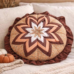 May include: A round, quilted throw pillow featuring a central star pattern made of folded fabric strips in shades of brown, peach, and white floral print. The pillow is finished with a gathered, dark brown ruffled edge around the entire perimeter.