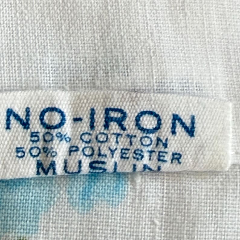 May include: Close-up of a white fabric label with the text "NO-IRON" in dark blue. Below, it reads "50% COTTON 50% POLYESTER MUSLIN". The fabric has a subtle, textured appearance.