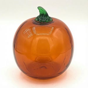 May include: A round, translucent orange glass pumpkin sculpture featuring a smooth, glossy surface and a textured, vibrant green glass stem attached to the top. The object reflects light, highlighting its spherical shape and clear glass construction.