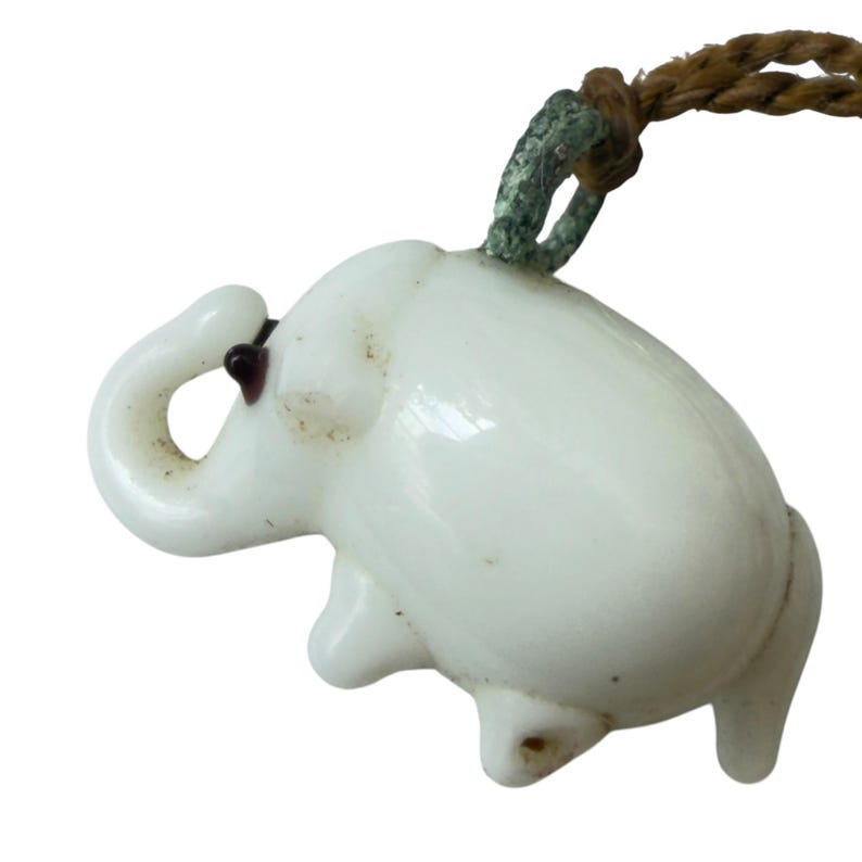 May include: A white elephant-shaped pendant made of smooth material, featuring a small dark eye detail and a curved trunk. The pendant is attached to a braided brown cord by a weathered green metal loop, creating a distinct contrast in texture and color.