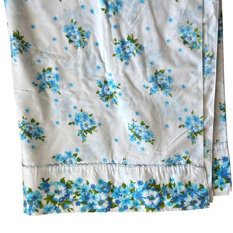 May include: White sheet set with a repeating pattern of blue floral bouquets and small blue star-shaped flowers. The bottom edge features a border of blue and green flowers. The fabric has a soft, slightly wrinkled appearance.