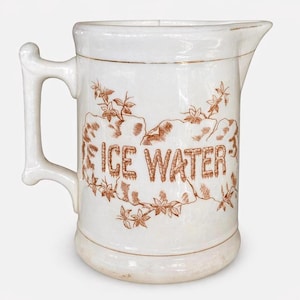 Könnte beinhalten: Weißer Keramikkrug mit Henkel und Ausguss. Der Krug hat braunen Text mit der Aufschrift "ICE WATER", umgeben von einem braunen Blumendesign. Eine dünne goldene Linie akzentuiert den oberen Rand.