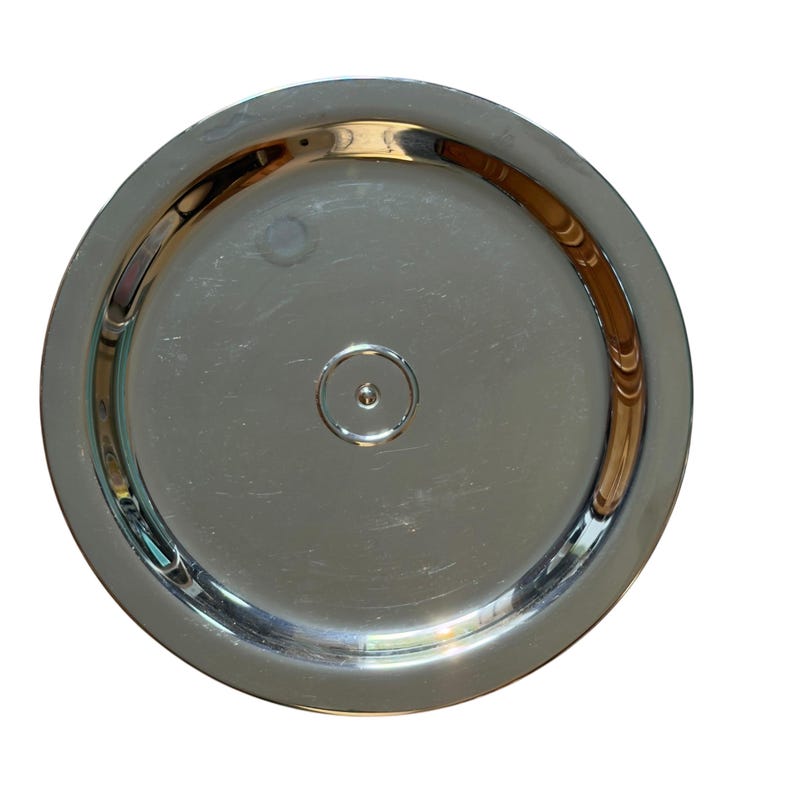 Op de afbeelding: Een cirkelvormig, gepolijst zilverkleurig metalen bord of dienblad met een glad, reflecterend oppervlak. Het object is voorzien van een verhoogde buitenrand en een kleine, gecentreerde cirkelvormige inkeping voor een strakke uitstraling.
