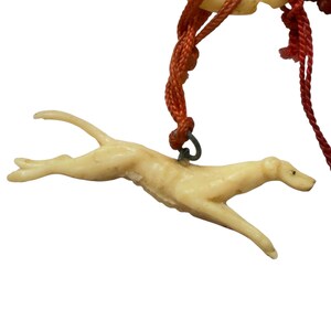 May include: A cream-colored, carved dog figurine in a running pose, suspended from a dark orange braided cord. The dog is depicted in mid-stride with its body elongated, featuring a small metal loop attached to its back for hanging purposes.