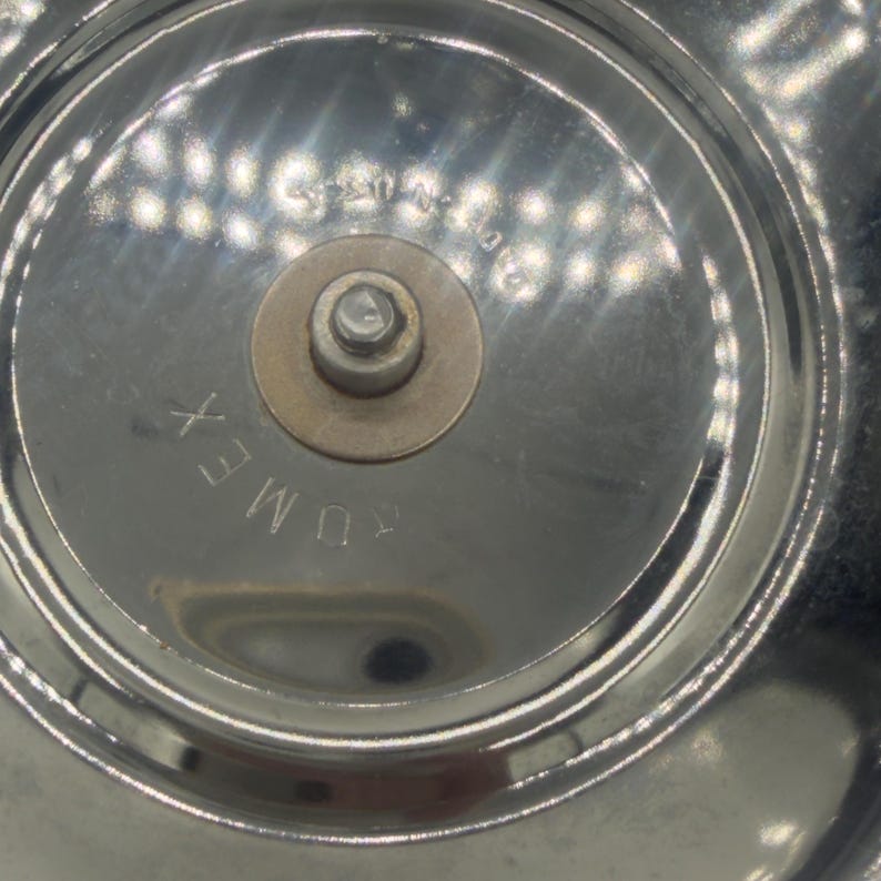 Op de afbeelding: Een cirkelvormig, reflecterend verchroomd metalen onderdeel met een centrale messingkleurige ring en een kleine uitstekende pin. Het woord "DUMEX" is in het oppervlak gestanst, samen met extra vage, gedeeltelijk zichtbare alfanumerieke tekst.