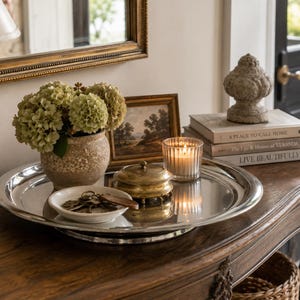 Op de afbeelding: Een grote ronde zilverkleurige metalen schaal met een beige keramische vaas met groene hortensia's, een klein wit schaaltje met metalen sleutels, een ronde koperen doos met deksel en een glazen waxinelichthouder met een brandende kaars.