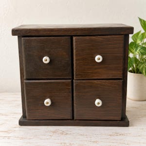 Könnte beinhalten: Eine dunkelbraune Aufbewahrungskommode aus Holz mit vier quadratischen Schubladen in einer Zwei-mal-zwei-Anordnung. Jede Schublade ist mit einem kleinen, runden, weißen Keramikknopf in der Mitte der Frontplatte versehen. Die Holzmaserung ist sichtbar.