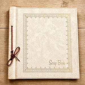 May include: Cream-colored scrapbook with a decorative gold border and the words "Scrap Book" in gold script. A brown cord ties the book closed. The scrapbook is on a wooden surface.