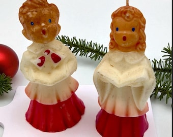 May include: Two vintage Christmas choir boy candles. The candles are shaped like children with orange hair, blue eyes, and red skirts. They are holding books and wearing white robes with red accents. The candles are on a white surface.