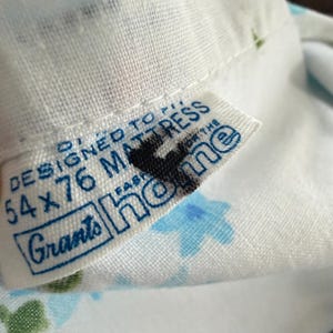 May include: Close-up of a white fabric with a blue floral pattern. A tag reads "DESIGNED TO FIT 54 x 76 MATTRESS" with the brand name "Grants home" in blue text. The fabric is slightly wrinkled.