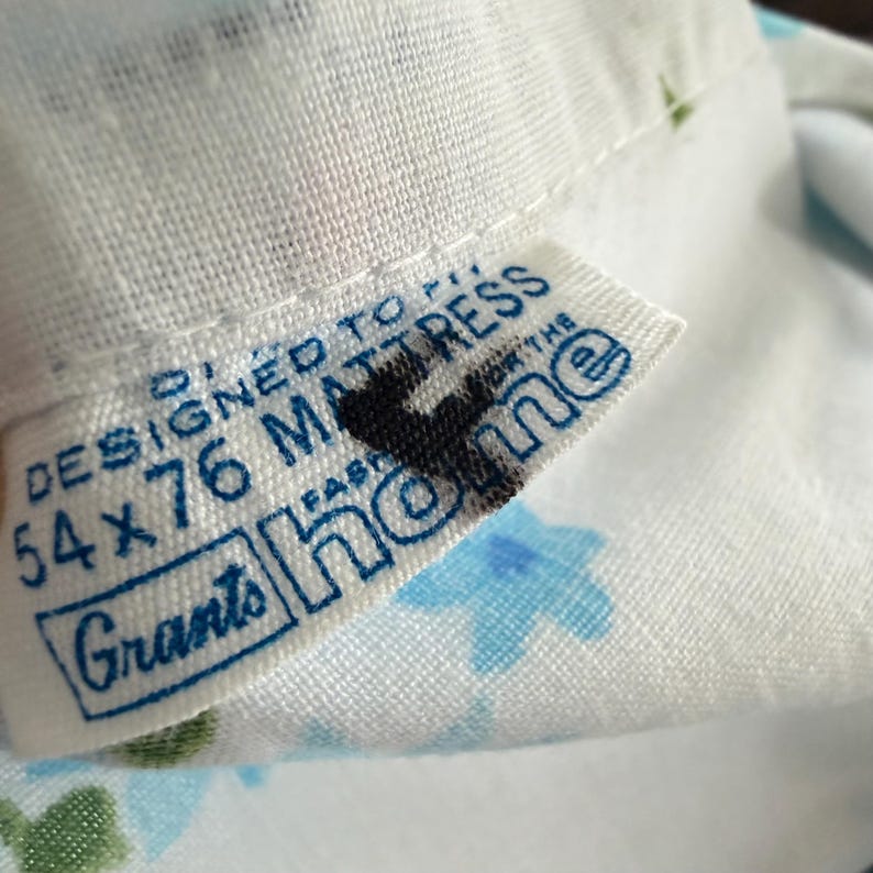 May include: Close-up of a white fabric with a blue floral pattern. A tag reads "DESIGNED TO FIT 54 x 76 MATTRESS" with the brand name "Grants home" in blue text. The fabric is slightly wrinkled.