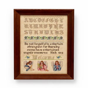 May include: A framed cross-stitch sampler in a brown wooden frame. The sampler displays the alphabet, a floral border, and a biblical quote. Below the quote are three angels and the word "Welcome".