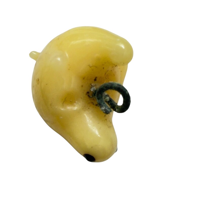May include: A pale yellow, smooth, rounded animal-shaped charm or pendant with a glossy finish. It features a small, dark, circular eye detail and a dark metal loop attached to the side for hanging, showing signs of oxidation on the metal surface.