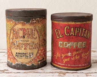 May include: Two vintage cylindrical metal coffee tins. The left tin is tan with a distressed Acme Brand Coffee label and red floral accents. The right tin is red with a gold El Capitan Coffee label and the text for your guest get the best.