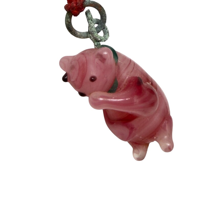 May include: A pink glass bear figurine charm with a dark green collar and small black eyes. The bear is suspended from a weathered, tarnished metal ring attached to a red cord. The glass has a translucent, swirled appearance with deeper pink accents.