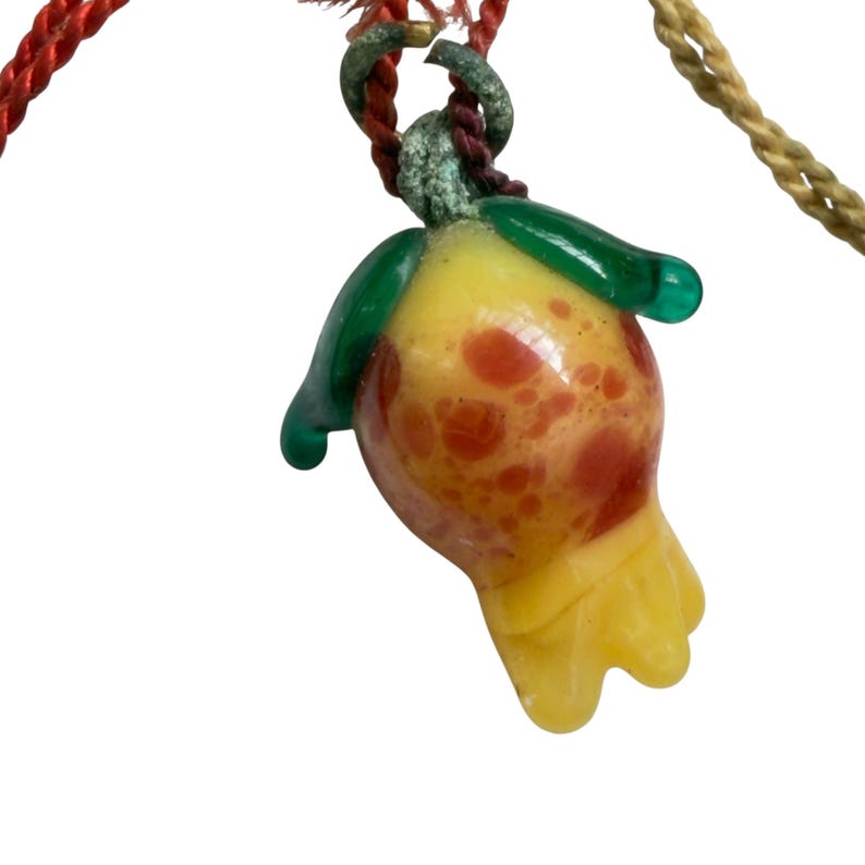 May include: A glass pendant shaped like a flower bud with a yellow and orange speckled body, green leaves at the top, and a yellow base. The pendant hangs from a red and gold twisted cord, featuring a small metal loop with visible oxidation.