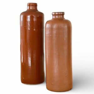 Puede incluir: Dos botellas antiguas de gres marrón con cuerpos cilíndricos y cuellos estrechos. Las botellas tienen un acabado brillante y se muestran sobre un fondo blanco. Las botellas son de diferentes tonos de marrón.