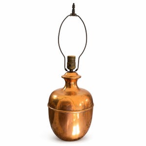 May include: A polished copper table lamp base with a rounded, bulbous body and a narrow neck. The lamp features a brass-coloured socket and a tall, oval-shaped metal harp attached to the top, designed to support a lampshade for lighting fixtures.