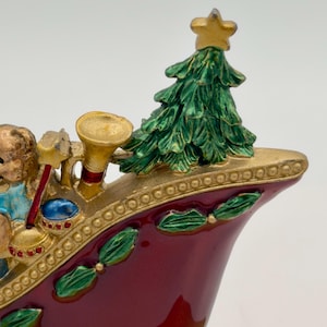 May include: A decorative sleigh figurine in deep red with gold trim, featuring a green Christmas tree with a gold star topper. The sleigh is adorned with green holly leaf accents and includes a small gold trumpet and a figure holding a red wand.